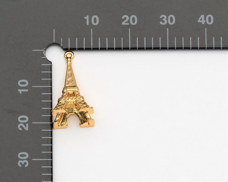 May include: Gold-coloured metal charm shaped like the Eiffel Tower. The charm has a loop at the top for attaching to a necklace or bracelet.