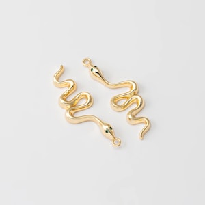 Peut inclure: Deux breloques dorées en forme de serpent avec des yeux en pierres vertes. Les breloques sont enroulées, avec une boucle en haut pour les attacher à des bijoux. Les serpents sont sur fond blanc.