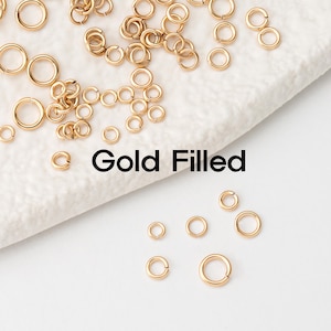 Puede incluir: Primer plano de un montón de anillos de salto rellenos de oro de varios tamaños. Los anillos de salto están esparcidos sobre una superficie blanca. El texto "Gold Filled" está escrito en letras negras debajo de los anillos de salto.