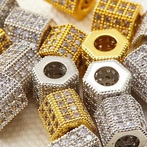 May include: Close-up of several hexagonal beads in gold and silver tones. The beads are encrusted with small, clear gemstones. The beads have a hole running through the center, and are likely used for jewelry making.