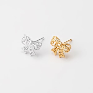 May include: Two bow-shaped stud earrings, one silver-colored and one gold-colored, are displayed against a white background. The bows have a delicate, openwork design with heart-shaped cutouts. The earrings are likely made of metal and are designed for pierced ears.
