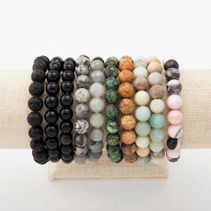 May include: A display of eight beaded bracelets in various colors and textures. The bracelets are made of round beads in shades of black, gray, green, brown, and pink. They are displayed on a beige fabric stand.