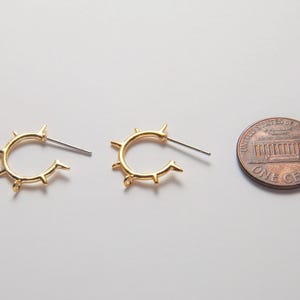 May include: A pair of gold-colored hoop earrings with a spiky design, next to a US one-cent coin for size comparison. The earrings have a post closure and are set against a white background.