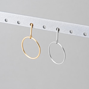 May include: Two minimalist hoop earrings, one gold-toned and one silver-toned, hang from a silver bar. The earrings feature a simple, circular design suspended from a straight post. The background is a soft, neutral gray.