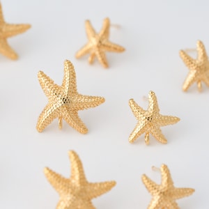 May include: Gold-colored starfish earrings with textured surfaces are scattered across a white background. The earrings are designed with five points, and each has a small loop at the base. The earrings are a beach-themed accessory.