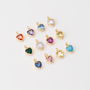 May include: Assortment of 12 gold heart-shaped charms with different colored gemstones. The charms are arranged in two rows on a white background.