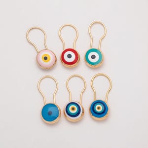 Könnte beinhalten: Sechs goldfarbene Schlüsselanhänger mit Emaille-Böser-Blick-Anhängern. Die Anhänger zeigen ein zentrales Augendesign in verschiedenen Farben, darunter Rosa, Rot, Blau und Türkis. Anordnung auf weißem Hintergrund.