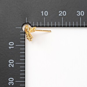 May include: A single gold-colored bow-shaped stud earring. The earring is highly detailed with a textured surface. The post is straight and thin. The earring is shown against a white background with a black ruler.