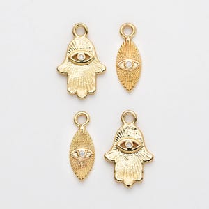 以下が含まれることがあります： 4つの金色のチャーム。2つはハムサハンドの形、2つは楕円形です。各チャームは、中央に小さな透明な石が付いた目のデザインが特徴です。チャームはテクスチャ加工された表面と、吊り下げ用のループが付いています。