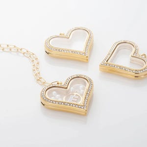 Könnte beinhalten: Goldfarbene herzförmige Medaillon-Halskette mit transparentem Mittelteil, umgeben von kleinen, funkelnden Kristallen. Das offene Medaillon zeigt ein perlenartiges Inneres und den Buchstaben "E". Zwei weitere herzförmige Medaillons sind ebenfalls vorhanden.