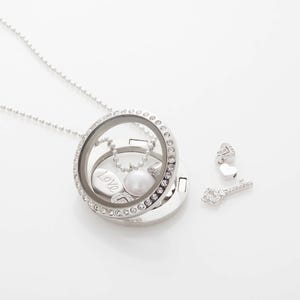 Puede incluir: Un collar con un medallón plateado y una cara de cristal transparente, incrustado con pequeños cristales transparentes. Dentro del medallón hay una perla, un dije con la palabra "Love" y una pequeña cadena. Los dijes adicionales incluyen un corazón y una llave.