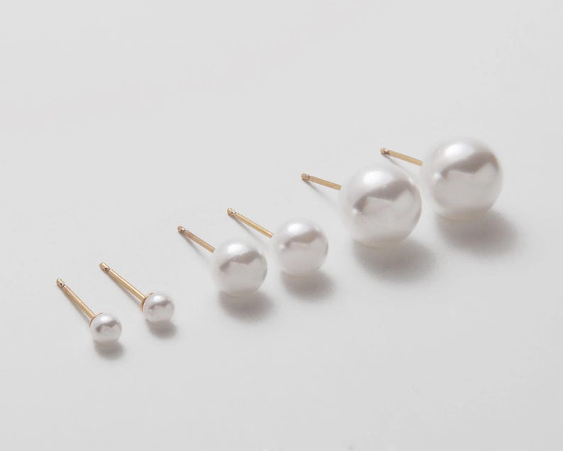 Op de afbeelding: Set van zes parel oorbel studs in verschillende maten, met gouden pinnen. De oorbel studs zijn gerangschikt in een lijn van de kleinste tot de grootste.