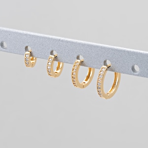 Könnte beinhalten: Vier goldfarbene Creolen in verschiedenen Größen, jeweils besetzt mit kleinen, klaren Edelsteinen. Die Ohrringe werden auf einer silbernen Stange präsentiert, die ihr Design und ihren Glanz hervorhebt. Schmuck.
