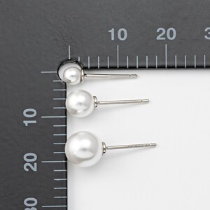 Op de afbeelding: Drie paar parel oorbelletjes in verschillende maten, gemeten met een liniaal. De oorbelletjes zijn zilverkleurig en hebben een witte parel.
