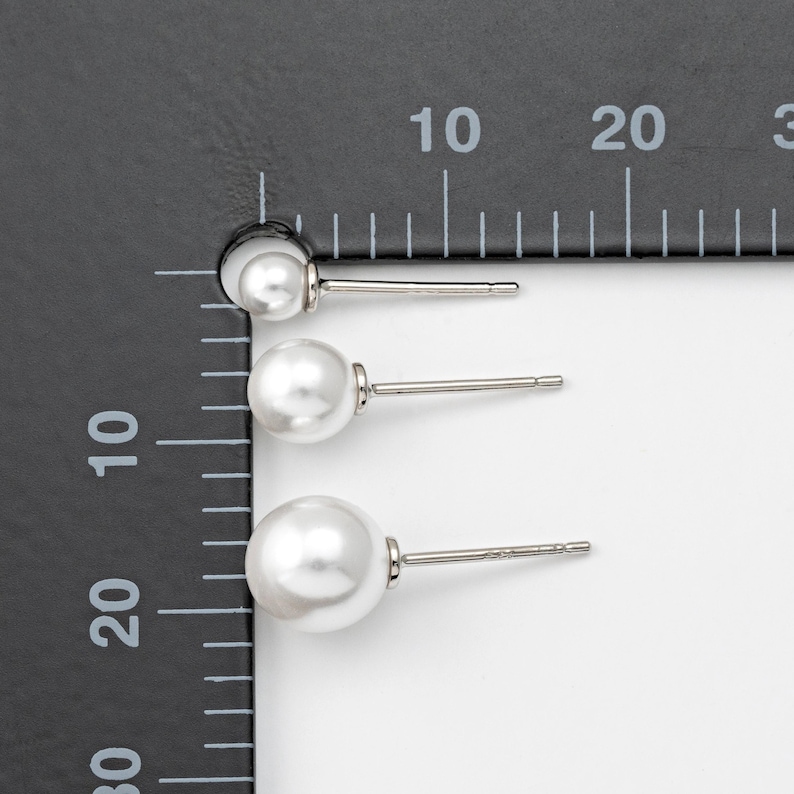 Op de afbeelding: Drie paar parel oorbelletjes in verschillende maten, gemeten met een liniaal. De oorbelletjes zijn zilverkleurig en hebben een witte parel.