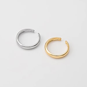 May include: Two ear cuffs, one silver and one gold, are displayed against a white background. The ear cuffs have a simple, minimalist design, with a smooth, rounded shape that curves around the ear. The silver cuff is on the left, and the gold cuff is on the right.