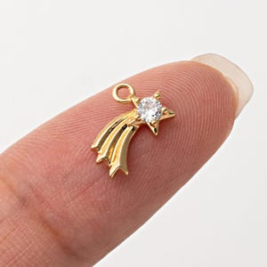May include: A gold-colored charm in the shape of a shooting star. The star has a clear, round gemstone in the center. The charm is small and detailed, with a loop at the top for attaching to a necklace or bracelet.