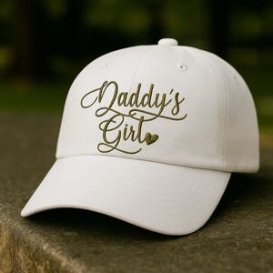 May include: White baseball cap with a curved brim. The cap features the embroidered text "Daddy's Girl" in a cursive font, with a small heart symbol. The hat is made of a soft fabric and has an adjustable strap.