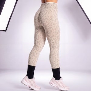 Peut inclure: Legging beige et marron clair à imprimé léopard. Le legging est porté avec des chaussettes noires et des chaussures de sport rose clair. Le legging a une taille haute et un motif texturé. Le modèle est sur une surface blanche.