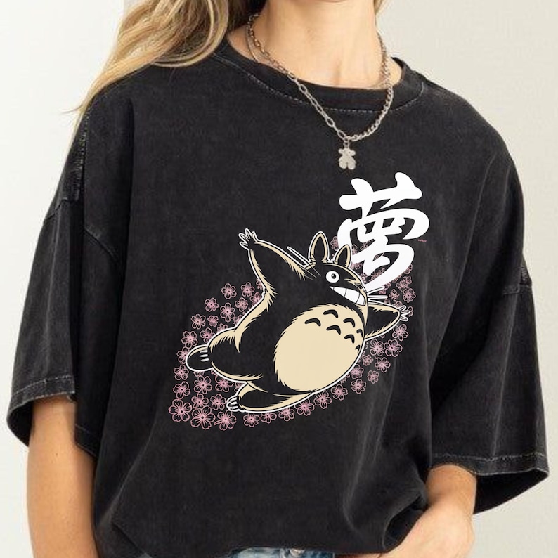 May include: Black t-shirt with a cartoon character design. The character has a cream-coloured belly and a smiling face, surrounded by pink flowers. The Japanese character for "dream" is printed above in white.