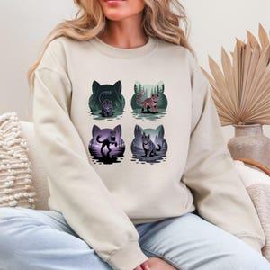 Może przedstawiać: Beżowa bluza z czterema ilustracjami z motywem kota. Każdy wzór przedstawia innego kota w innym otoczeniu, takim jak las i odbijający wodę zbiornik. Ilustracje wykorzystują odcienie zieleni, fioletu i różu. Bluza jest w neutralnym kolorze.