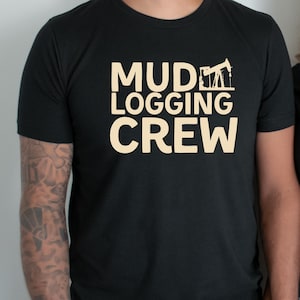 Op de afbeelding: Zwart T-shirt met ronde hals met de woorden "MUD LOGGING CREW" in lichtbruin gedrukt. Het ontwerp bevat een illustratie van een boortoren. Het shirt is gemaakt van een zacht materiaal.