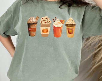 May include: Sage green t-shirt featuring a graphic of four different fall-themed coffee drinks. The drinks include a pumpkin spice latte, a coffee with heart-shaped sprinkles, a drink with whipped cream and a leaf, and a coffee with a pumpkin.