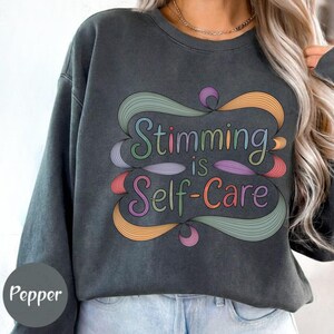 May include: Dark gray sweatshirt with the text "Stimming is Self-Care" in colorful, swirling script. The design features shades of green, orange, pink, and purple. The sweatshirt is a relaxed fit.