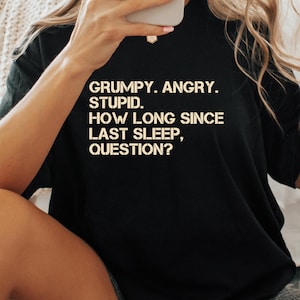 Op de afbeelding: Zwart T-shirt met witte tekst: "GRUMPY. ANGRY. STUPID. HOW LONG SINCE LAST SLEEP, QUESTION?" Het shirt is een casual item, geschikt voor dagelijks gebruik. De tekst is in een vet, schreefloos lettertype.