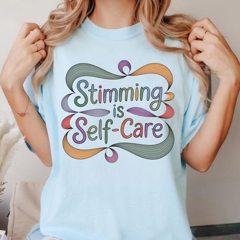 May include: Light blue t-shirt with the text "Stimming is Self-Care" in a colorful, stylized design. The text is in green, purple, orange, and red, with decorative swirls. The shirt is a short-sleeved crew neck.