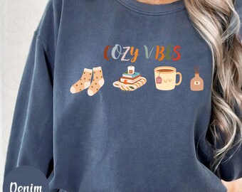 Könnte beinhalten: Denimblaues Sweatshirt mit dem Schriftzug "COZY VIBES" in bunten Buchstaben. Die Grafik zeigt Socken, Bücher, eine Tasse Tee und einen Diffusor. Das Sweatshirt hat eine lockere Passform.