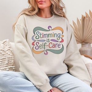 May include: Beige sweatshirt with the text "Stimming is Self-Care" in a colorful, stylized design. The text is framed by a decorative, swirling border with shades of green, orange, and purple. The sweatshirt is a neutral color.
