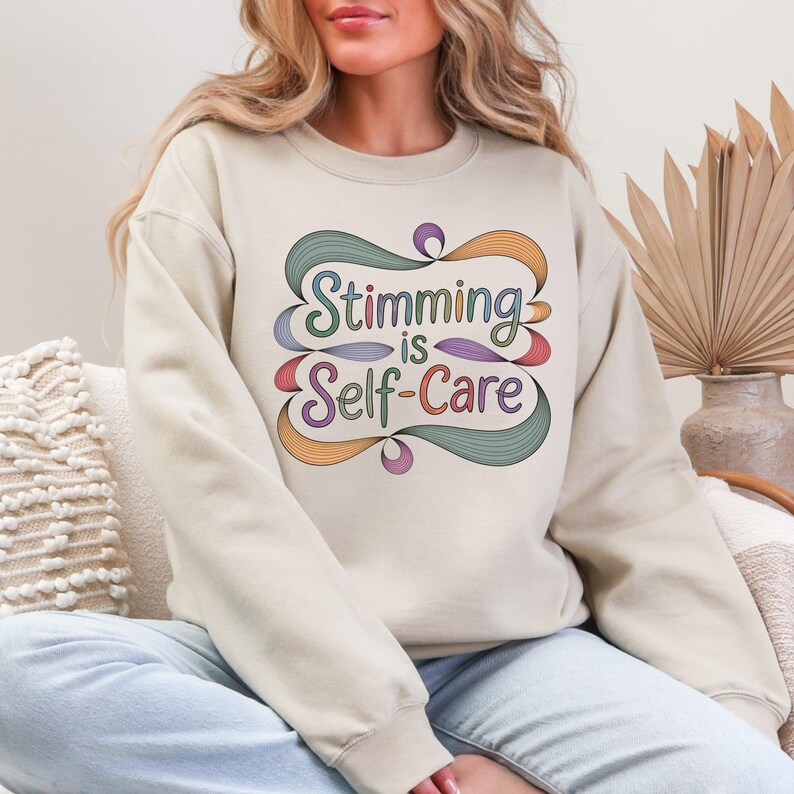 May include: Beige sweatshirt with the text "Stimming is Self-Care" in a colorful, stylized design. The text is framed by a decorative, swirling border with shades of green, orange, and purple. The sweatshirt is a neutral color.