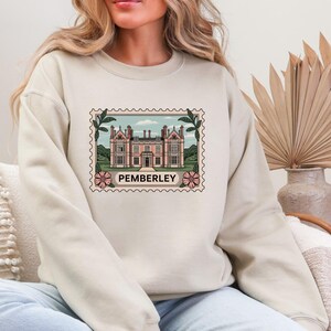 Peut inclure: Un sweat-shirt crème avec un motif de timbre-poste représentant un bâtiment rose avec l'inscription "PEMBERLEY". Le timbre est encadré de feuilles vertes et de fleurs roses. Le sweat-shirt est de style décontracté et confortable.