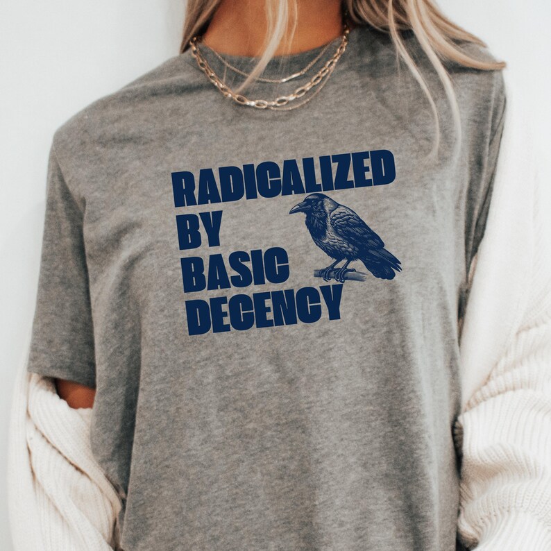 May include: A gray t-shirt with the text "RADICALIZED BY BASIC DECENCY" in navy blue. A navy blue illustration of a crow perched on a branch is to the right of the text. The shirt has short sleeves.