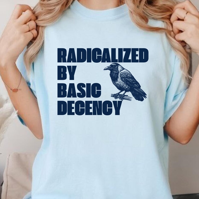 May include: Light blue t-shirt with the text "RADICALIZED BY BASIC DECENCY" in dark blue block letters. A detailed illustration of a crow perched on a branch is to the right of the text. The shirt has short sleeves.