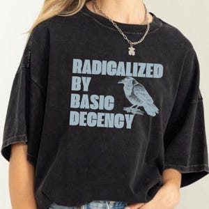 May include: Black t-shirt with the text "RADICALIZED BY BASIC DECENCY" in light blue block letters. A light blue illustration of a raven perched on a branch is to the right of the text. The shirt has a crew neck and short sleeves.