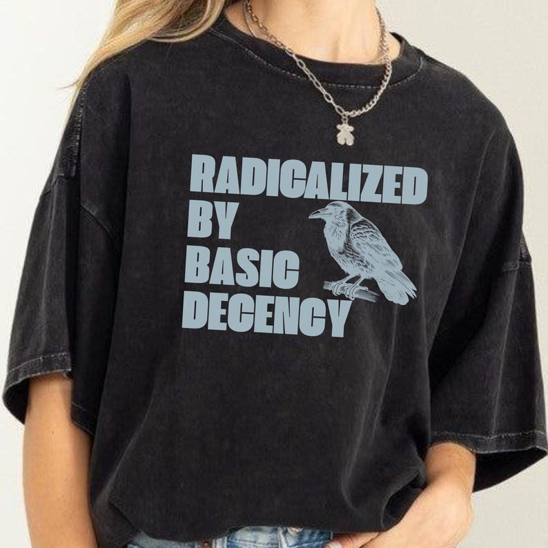 May include: Black t-shirt with the text "RADICALIZED BY BASIC DECENCY" in light blue block letters. A light blue illustration of a raven perched on a branch is to the right of the text. The shirt has a crew neck and short sleeves.