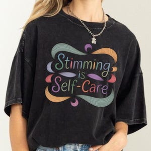 May include: Black t-shirt with the text "Stimming is Self-Care" in a colorful, stylized design. The design features swirling shapes in shades of green, orange, pink, and purple. The shirt has a relaxed fit and short sleeves.