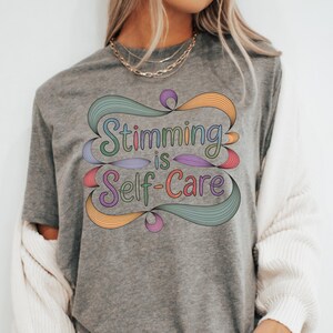 May include: A gray t-shirt with the text "Stimming is Self-Care" in colorful, stylized lettering. The design features swirling ribbons in shades of blue, green, orange, and purple. The shirt is a casual, everyday wear item.