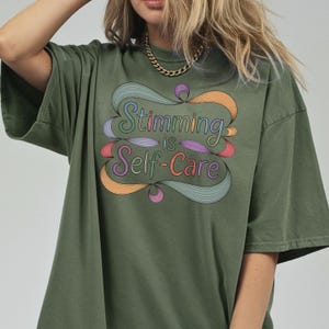 May include: Olive green t-shirt with the text "Stimming is Self-Care" in a colorful, stylized design. The design features swirling lines in shades of blue, orange, purple, and pink. The shirt has short sleeves and a relaxed fit.
