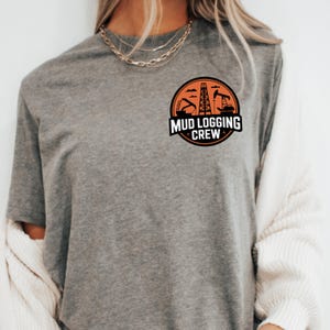 Op de afbeelding: Grijs t-shirt met een cirkelvormig ontwerp met de tekst "MUD LOGGING CREW". Het ontwerp toont zwarte en oranje silhouetten van olieboortorens en industriële machines.