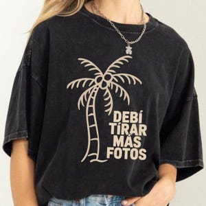 May include: Black t-shirt with a beige palm tree graphic and the Spanish text "Debí Tirar Más Fotos." The design features a detailed palm tree illustration. The shirt has a relaxed fit and short sleeves. The fabric has a washed-out effect.
