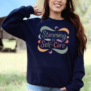 May include: Navy blue sweatshirt with the text "Stimming is Self-Care" in a colorful, swirling design. The text is in a variety of colors, including green, orange, pink, and purple. The sweatshirt is a long-sleeved crew neck.