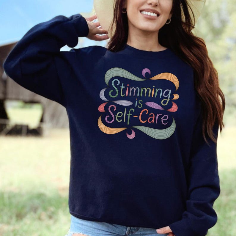 May include: Navy blue sweatshirt with the text "Stimming is Self-Care" in a colorful, swirling design. The text is in a variety of colors, including green, orange, pink, and purple. The sweatshirt is a long-sleeved crew neck.