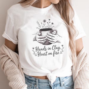 May include: White t-shirt with a black and white illustration of hands holding a mug with flowers. The text "Hands in Clay, Heart on fire" is below the illustration. The shirt is worn by a person.