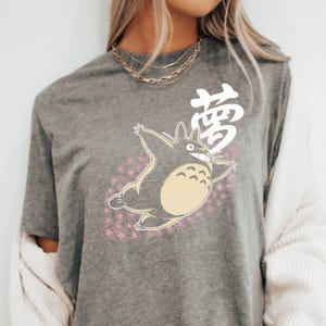 May include: Grey t-shirt with a cartoon character, cream-coloured belly, and smiling face, surrounded by pink flowers. The Japanese character for "dream" is printed above the character.