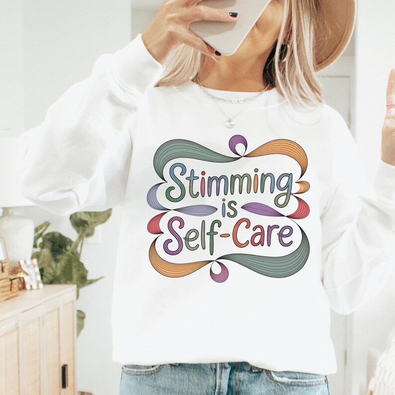 May include: White sweatshirt with the text "Stimming is Self-Care" in a colorful, stylized design. The text is in a whimsical font, with the words outlined in various colors including green, purple, orange, and red. The sweatshirt has long sleeves.