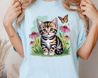 May include: Light blue t-shirt featuring a cartoon kitten with black and orange stripes, green eyes, and a butterfly. The kitten is surrounded by pink flowers and green grass. The shirt is a solid light blue color.