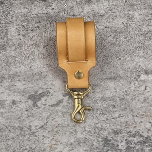 May include: A tan leather key fob with a brass-colored swivel clasp and a brass rivet. The key fob has a loop for attaching to a belt or bag. The leather is a light brown color.